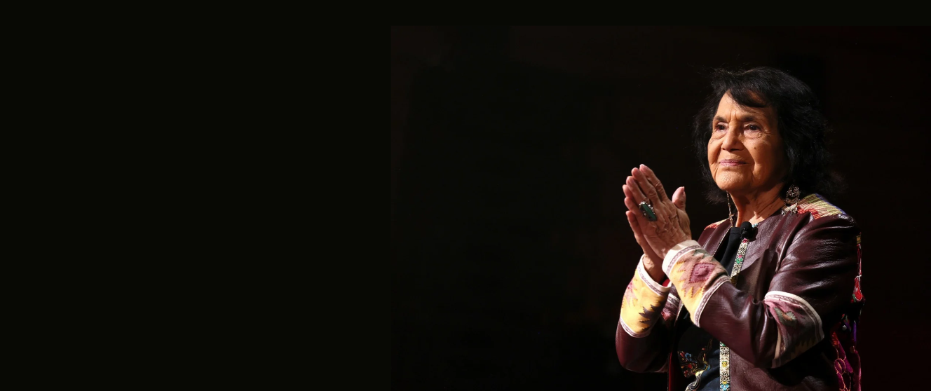 A photograph shows Huerta with her hands together, wearing a burgundy jacket with colorful embroidered patterns against a black background.