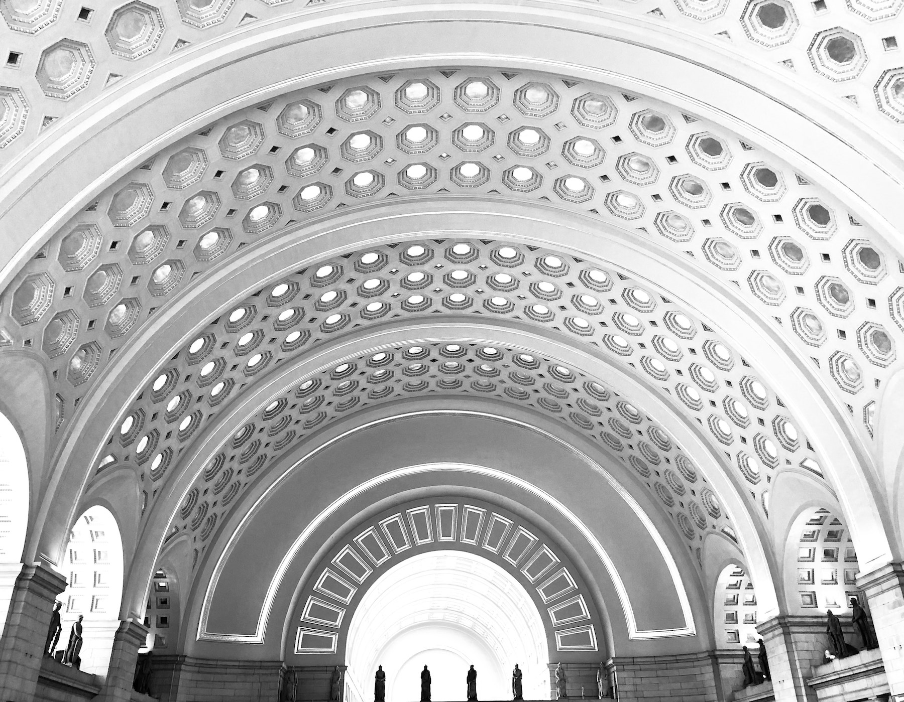 Black and white interior of Union Station in Washington, DC. The ceiling is vast, arching and ornate. Statues stand tall below an arch way.